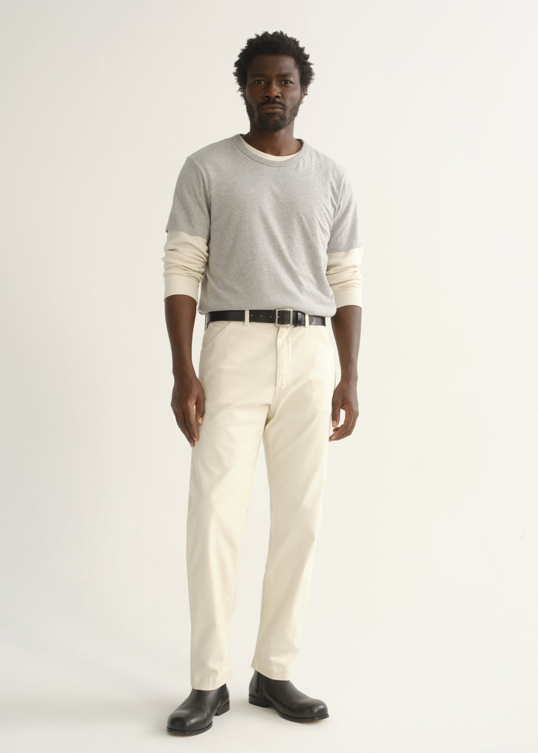 Person wearing a light grey crewneck t-shirt with cream trousers and black shoes, front full body view on a light grey background.