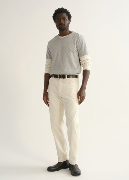 Person wearing a light grey crewneck t-shirt with cream trousers and black shoes, angled front full body view on a light grey background.