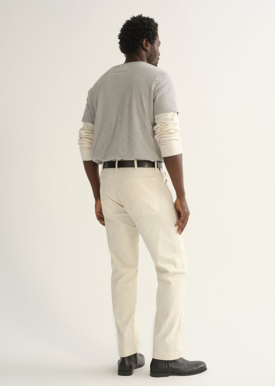 Person wearing a light grey crewneck t-shirt with cream trousers and black shoes, back full body view on a light grey background.