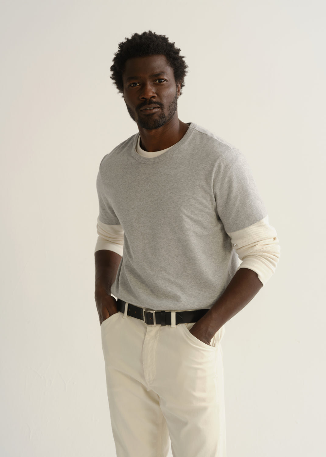 Person wearing a light grey crewneck t-shirt with cream trousers and a black belt, front cropped view on a light grey background.