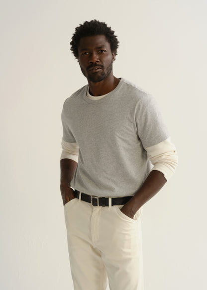 Person wearing a light grey crewneck t-shirt with cream trousers and a black belt, front cropped view on a light grey background.