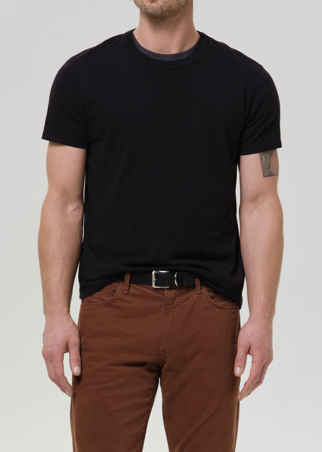 A man wearing a plain black short-sleeve crewneck t-shirt tucked into brown pants with a black belt, photographed against a grey background.