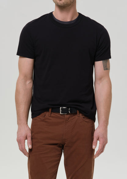 A man wearing a plain black short-sleeve crewneck t-shirt tucked into brown pants with a black belt, photographed against a grey background.