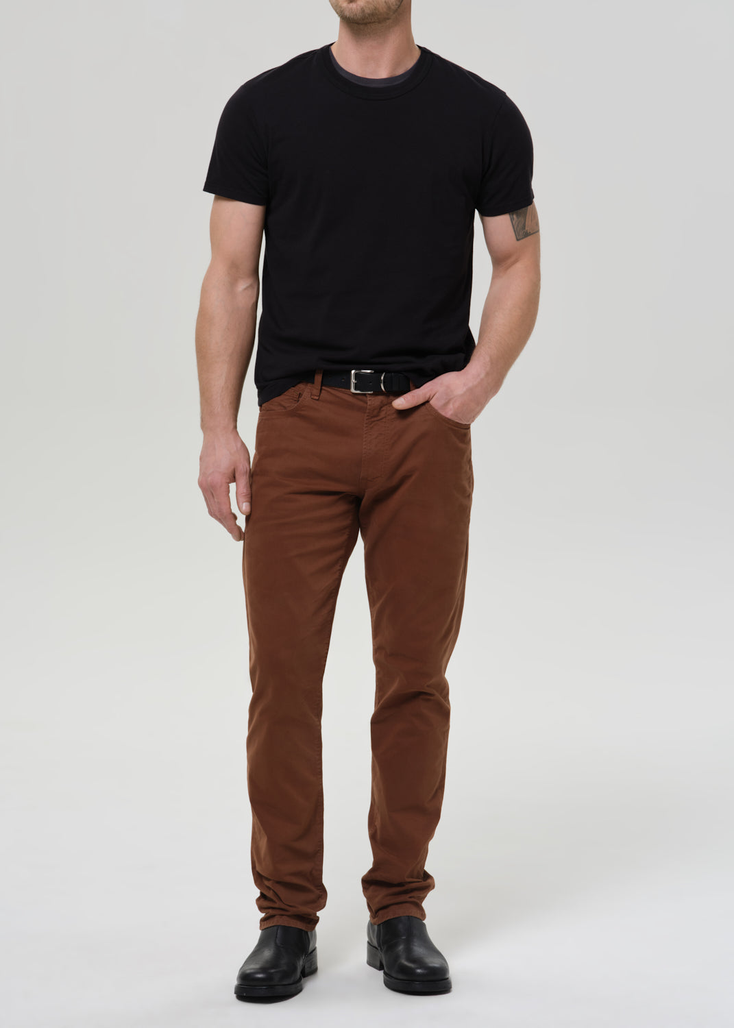 A man modeling a black crewneck t-shirt and brown slim-fit pants with a black belt and black leather shoes, standing against a grey background.