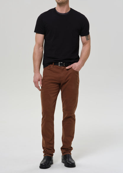 A man modeling a black crewneck t-shirt and brown slim-fit pants with a black belt and black leather shoes, standing against a grey background.