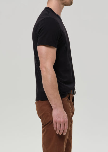 A man shown in side profile wearing a black short-sleeve crew-neck t-shirt and brown pants, photographed against a grey background.
