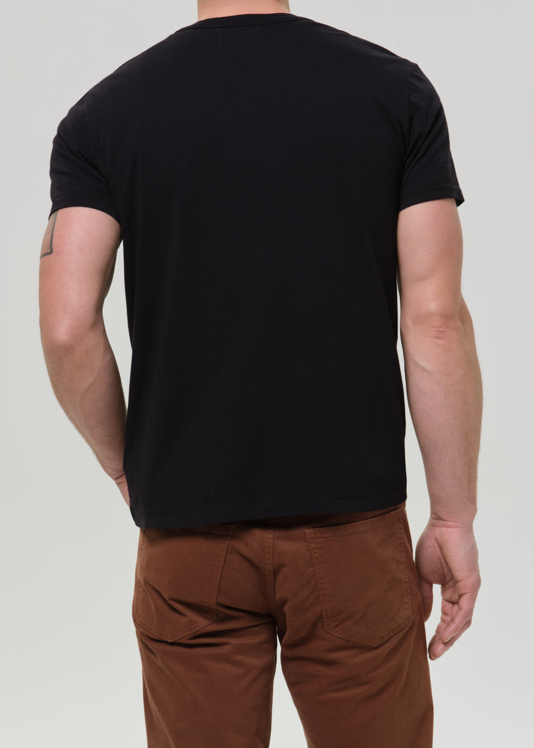 A man viewed from the back wearing a plain black short-sleeve crewneck t-shirt and brown pants, against a grey background.