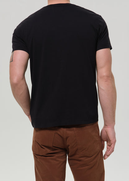 A man viewed from the back wearing a plain black short-sleeve crewneck t-shirt and brown pants, against a grey background.