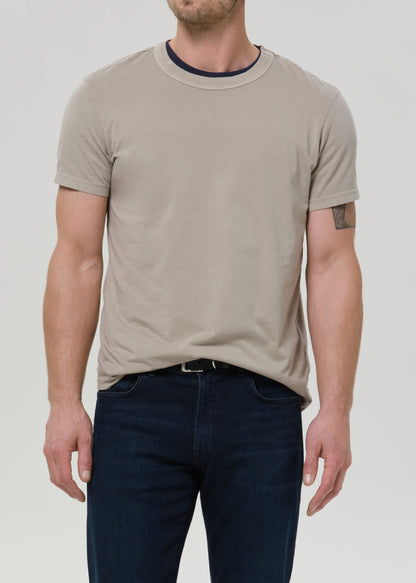 Man wearing a beige t-shirt and dark jeans on a plain background
