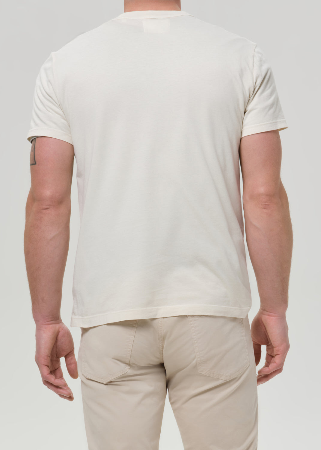 Person wearing a cream crewneck short sleeve tee with light beige straight leg pants viewed from the back on a grey background.