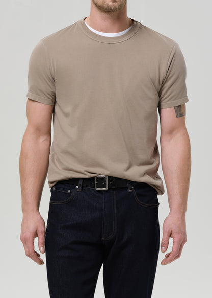 Man wearing a beige t-shirt and dark jeans on a plain background