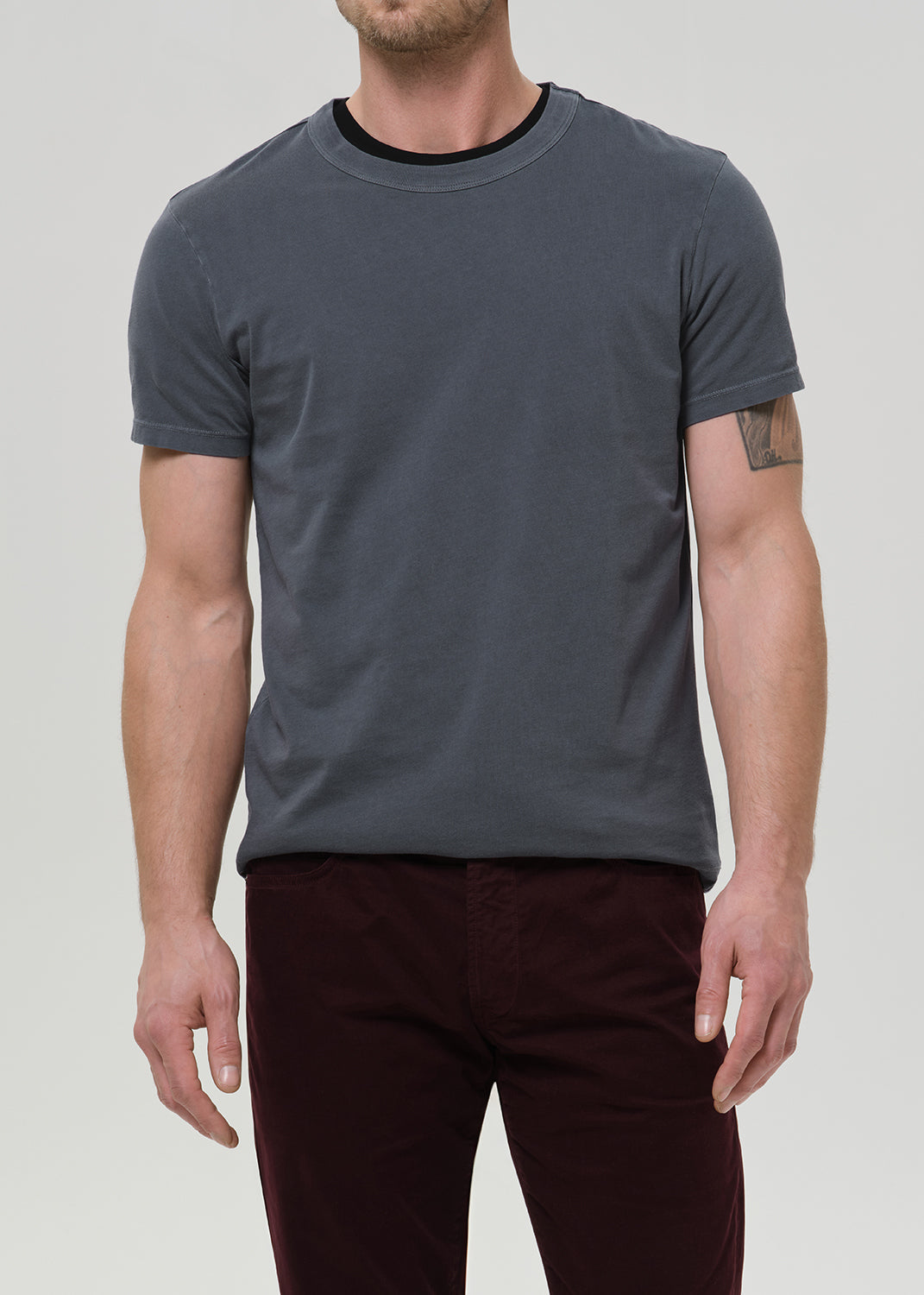 A man modeling a dark gray crewneck t-shirt and dark burgundy corduroy trousers, paired with black leather boots, against a plain light gray studio background.