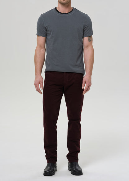 A man modeling a dark gray crewneck t-shirt and dark burgundy corduroy trousers, paired with black leather boots, against a plain light gray studio background.