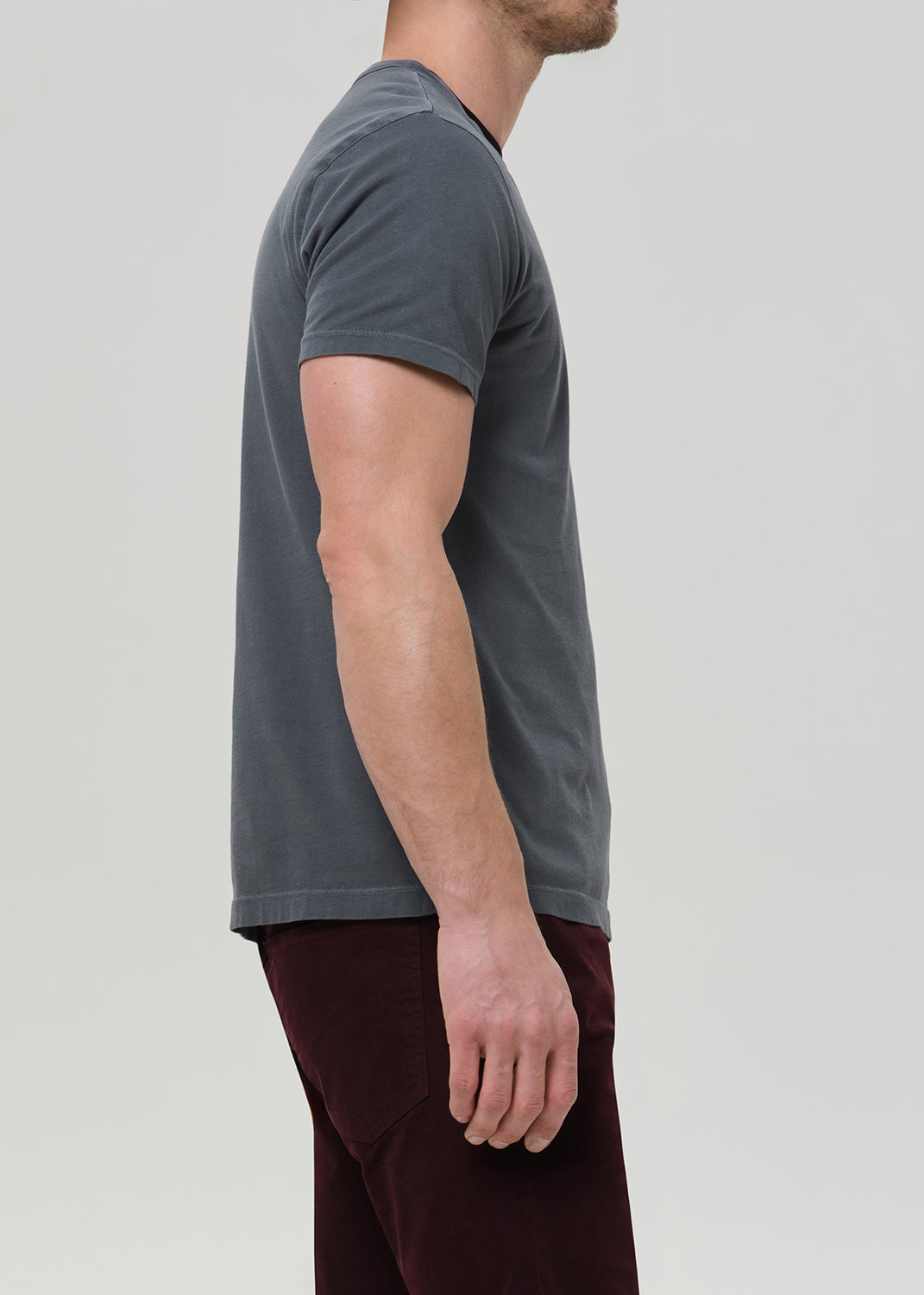 A man modeling a dark gray crewneck t-shirt and dark burgundy corduroy trousers, paired with black leather boots, against a plain light gray studio background.