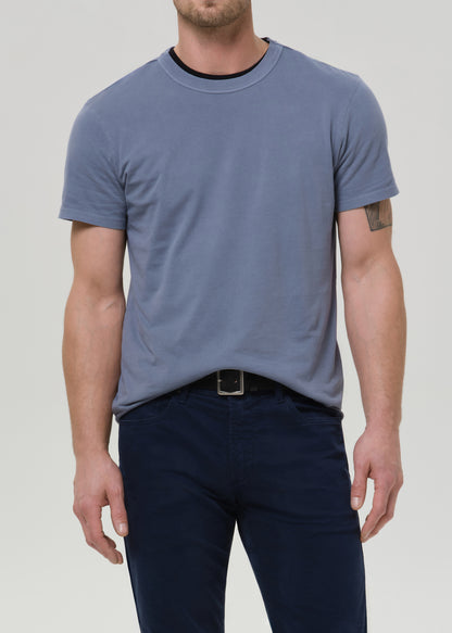 A man wearing a slate blue crewneck t-shirt with a black ribbed collar trim, paired with dark navy pants, shown from the chin down against a grey background.