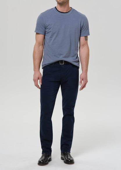 A man wearing a slate blue crewneck t-shirt, dark navy blue pants, a black belt, and black leather shoes, standing against a grey background.