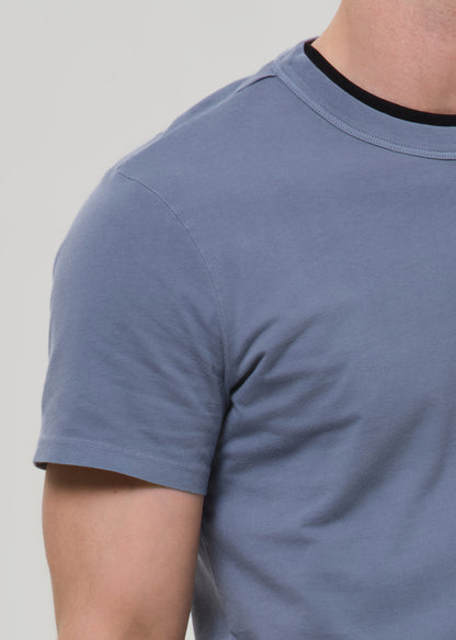 Close-up of a person wearing a short-sleeve crew-neck t-shirt in dusty blue with a black ribbed collar trim, against a grey background.