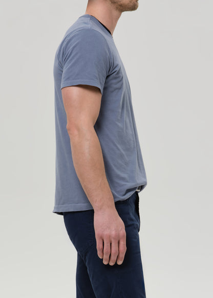 A man shown in profile wearing a muted slate blue short-sleeve crewneck t-shirt and dark navy pants, photographed against a grey background.
