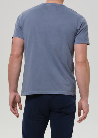 A man viewed from the back wearing a muted slate blue crewneck t-shirt and dark navy pants, set against a grey background.