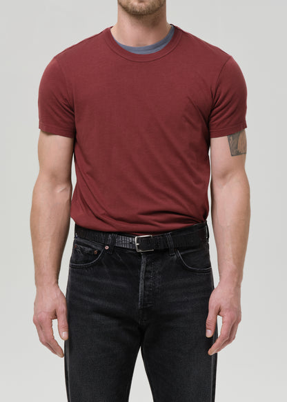 Man wearing a maroon t-shirt and black jeans against a grey background