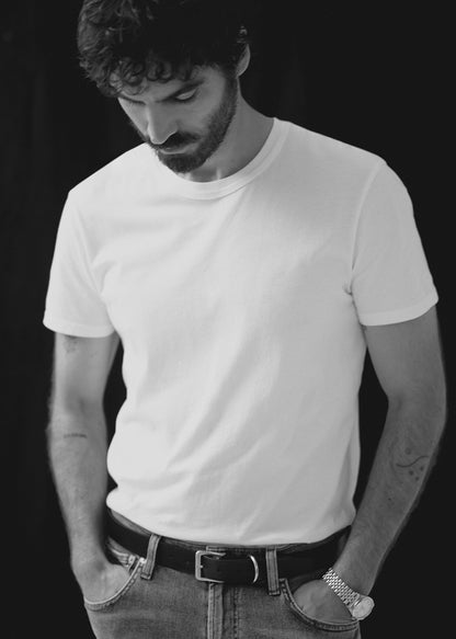 A man wearing a plain white short-sleeve crewneck t-shirt, styled with a black belt and jeans, photographed in black and white against a dark background.
