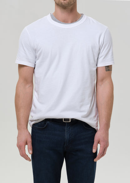 A man wearing a plain white short-sleeve crewneck t-shirt and dark blue jeans, shown from the chin down against a grey background.