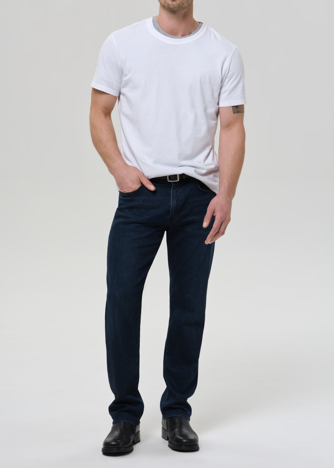 Man wearing a white t-shirt and dark jeans on a grey background.