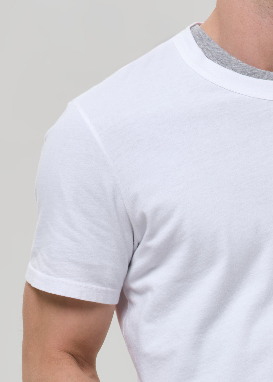 Close-up of a person wearing a plain white short-sleeve crew-neck t-shirt with a visible light gray inner neck trim, against a grey background.