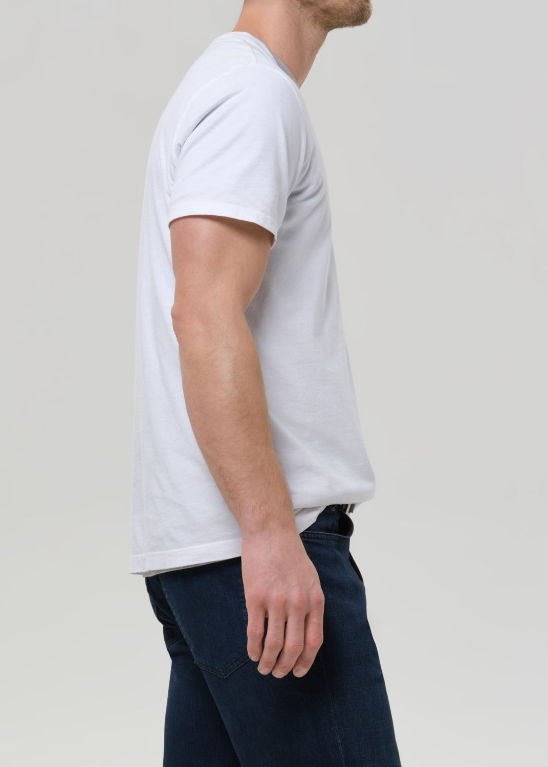 A man shown from the side wearing a plain white short-sleeve crew-neck t-shirt and dark blue jeans, photographed against a grey background.