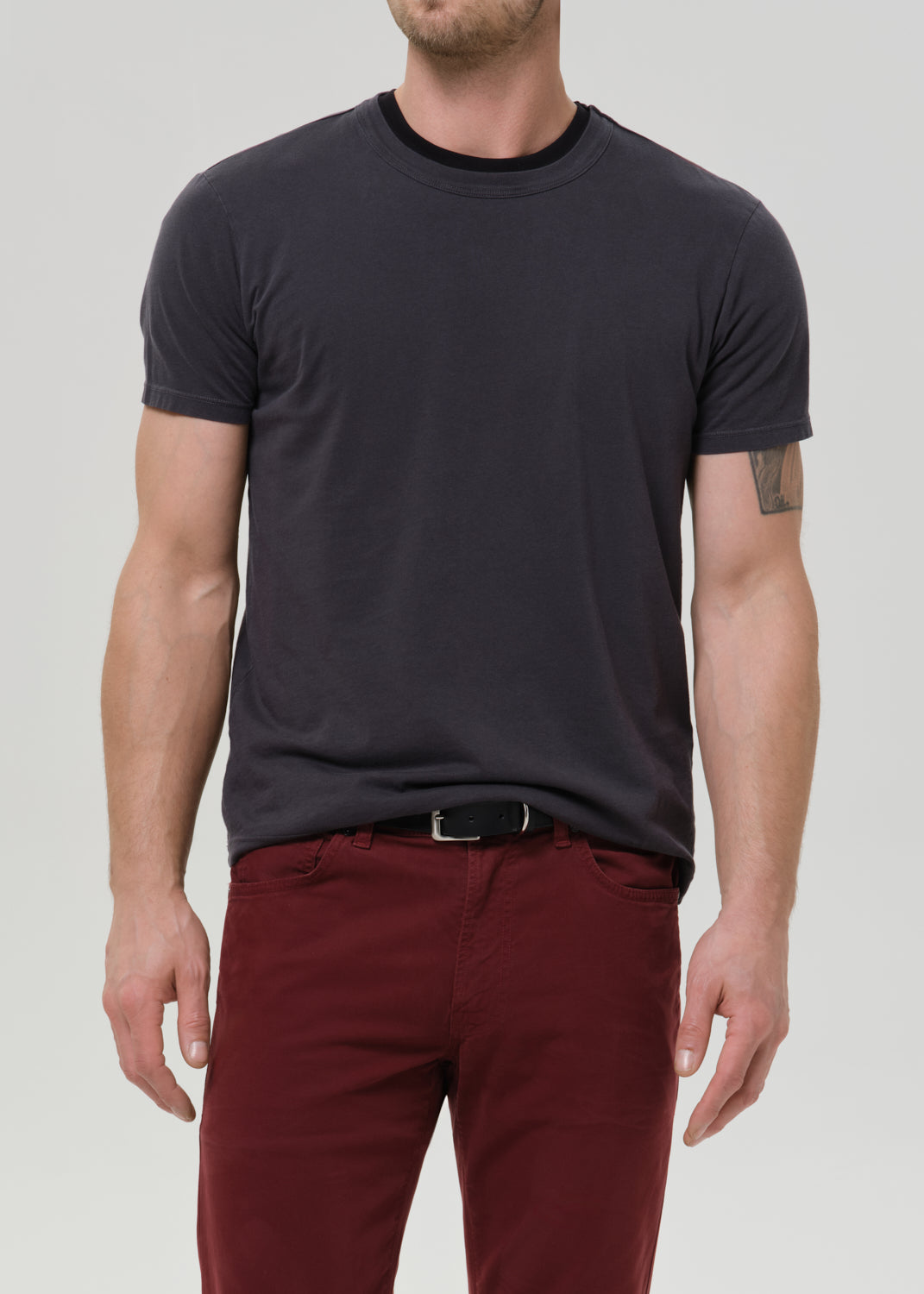 A man wearing a dark charcoal grey crew-neck t-shirt and burgundy pants, shown from the chin down against a grey background.