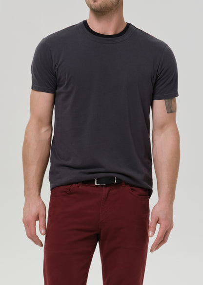 A man wearing a dark charcoal grey crew-neck t-shirt and burgundy pants, shown from the chin down against a grey background.