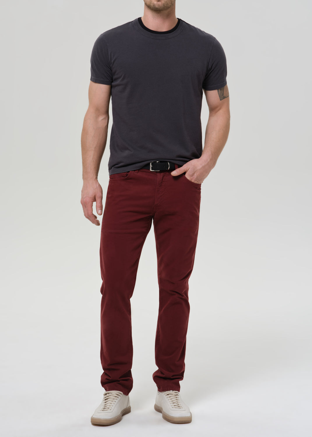 A man wearing slim-fit burgundy trousers, a dark charcoal grey t-shirt, a black belt, and off-white sneakers, standing against a grey background.