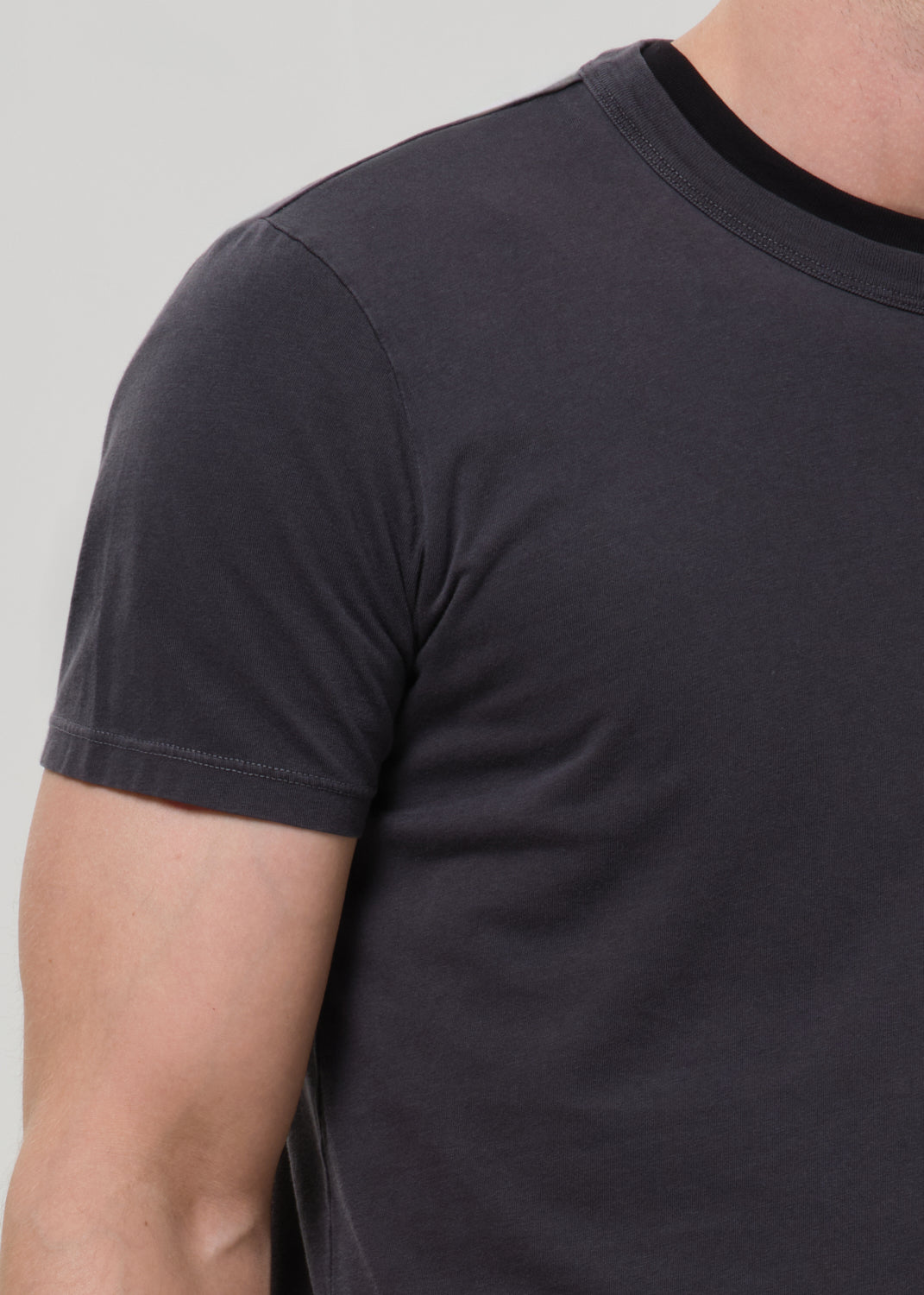 Close-up of a person wearing a dark charcoal grey short-sleeve crewneck t-shirt with a contrasting black ribbed collar, shown against a grey background.