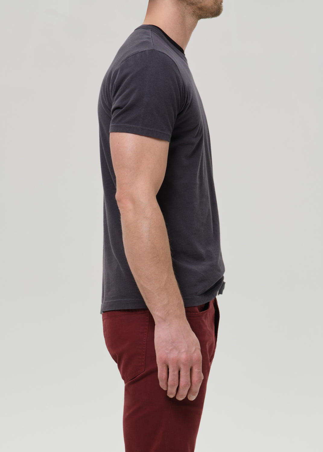 A man shown in side profile wearing a dark charcoal grey short-sleeve crewneck t-shirt and burgundy pants, photographed against a grey background.