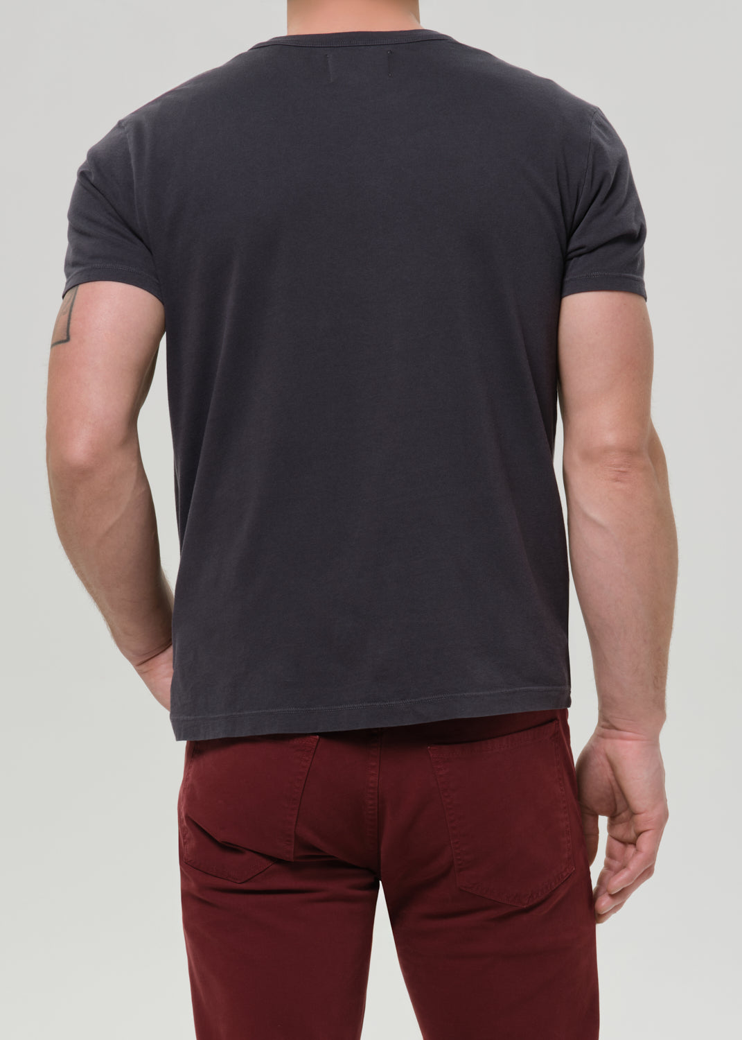 A man shown from the back wearing a dark charcoal grey short-sleeve t-shirt and burgundy pants, against a grey background.