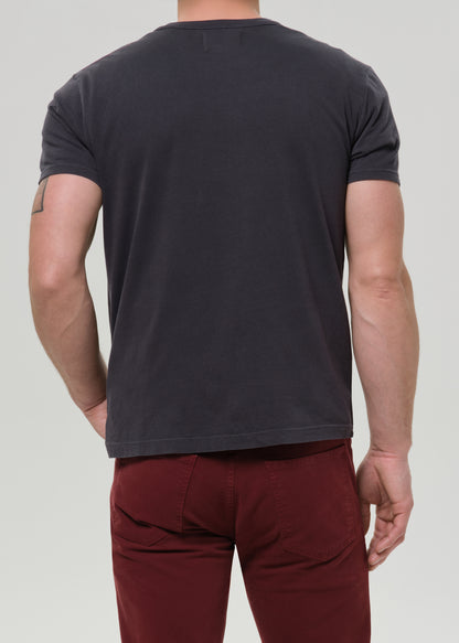 A man shown from the back wearing a dark charcoal grey short-sleeve t-shirt and burgundy pants, against a grey background.