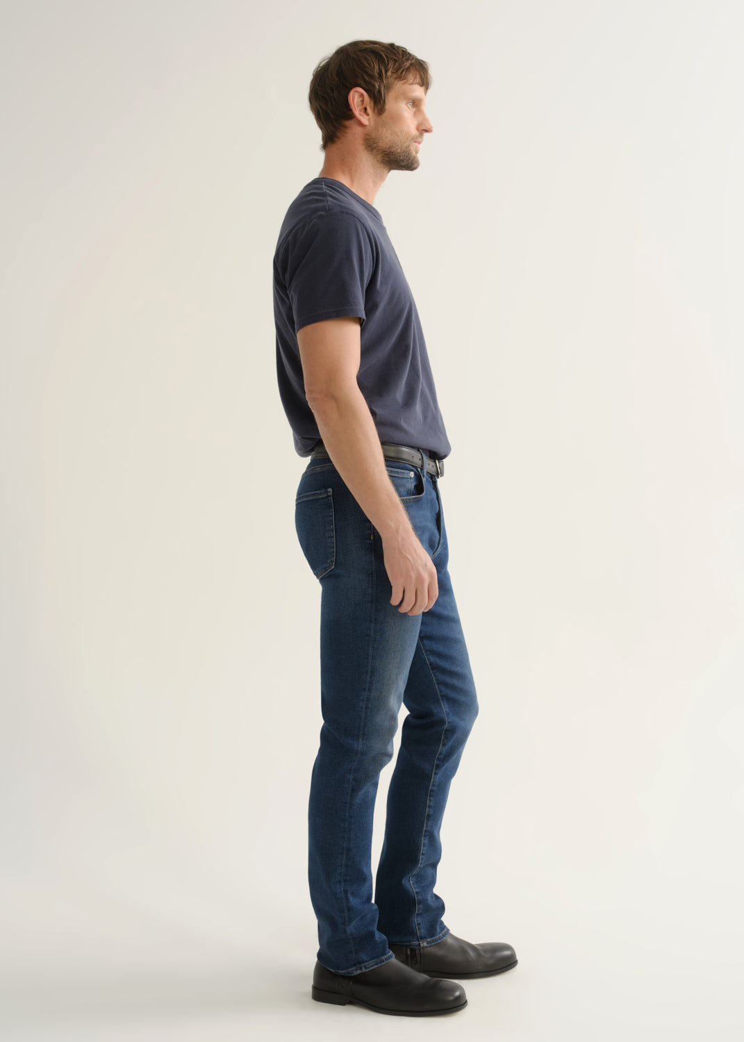 Person wearing a dark charcoal short sleeve crewneck t-shirt with blue jeans and black shoes, side full body view on a light grey background.