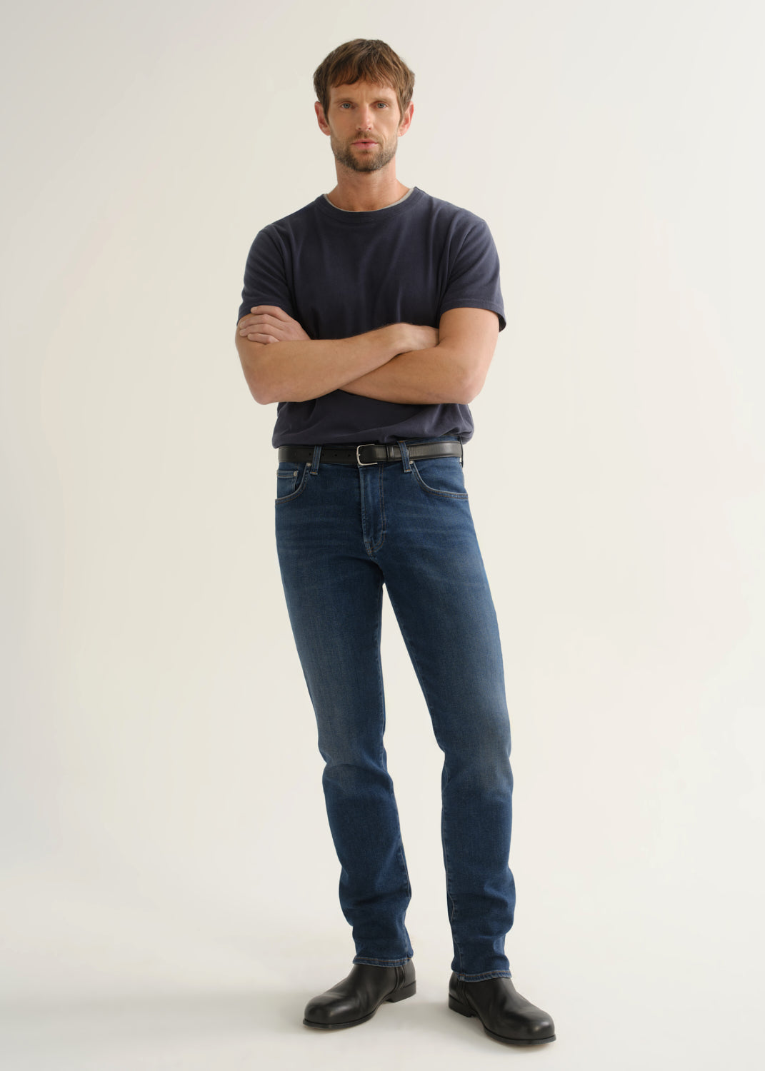 Person wearing a dark charcoal short sleeve crewneck t-shirt with blue jeans and black shoes, front full body view on a light grey background.