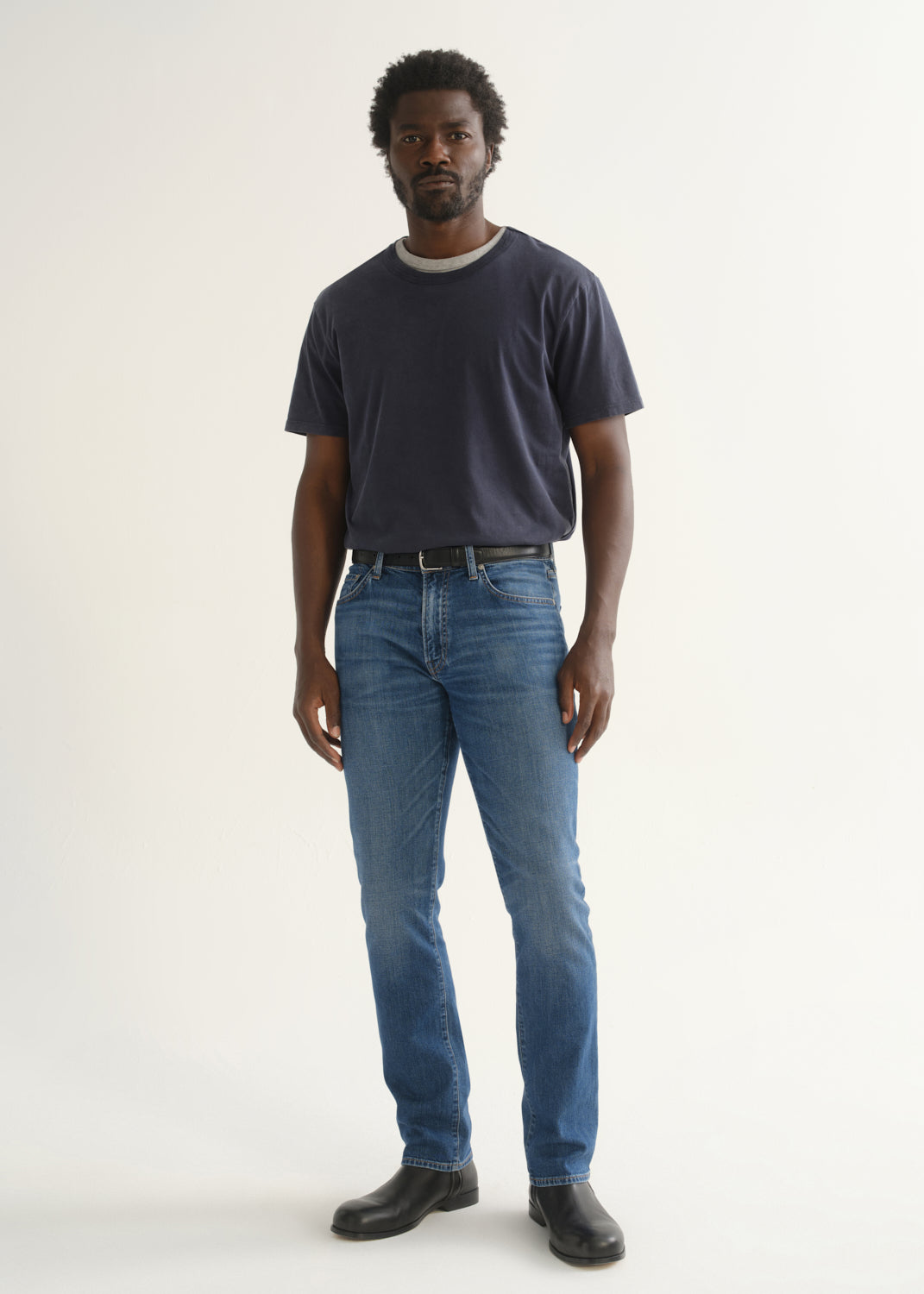 Person wearing a navy blue short sleeve crewneck t-shirt with medium blue jeans and black shoes, front full body view on a light grey background.