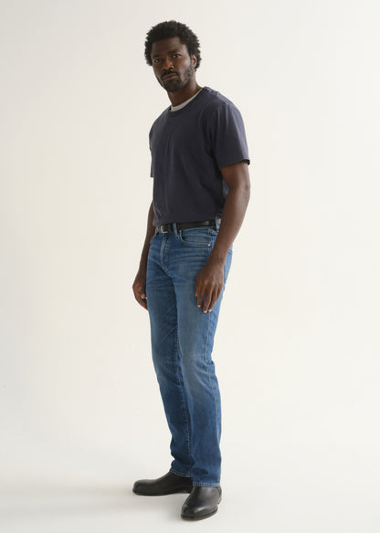 Person wearing a navy blue short sleeve crewneck t-shirt with medium blue jeans and black shoes, angled front full body view on a light grey background.