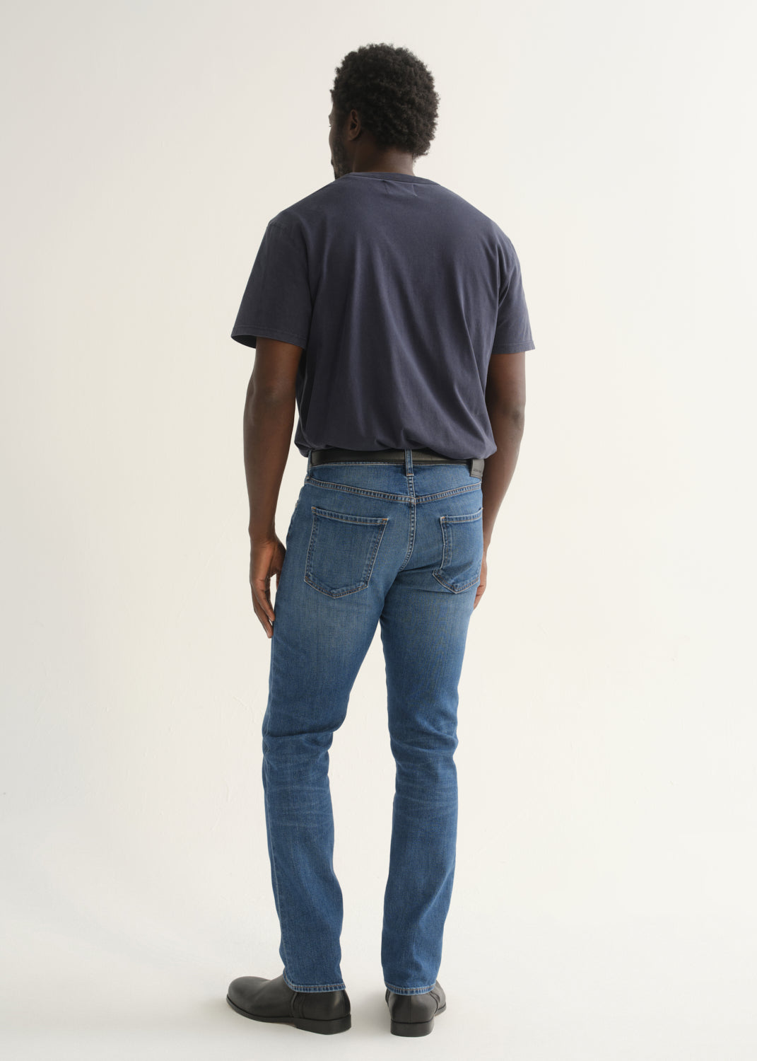 Person wearing a navy blue short sleeve crewneck t-shirt with medium blue jeans and black shoes, back full body view on a light grey background.