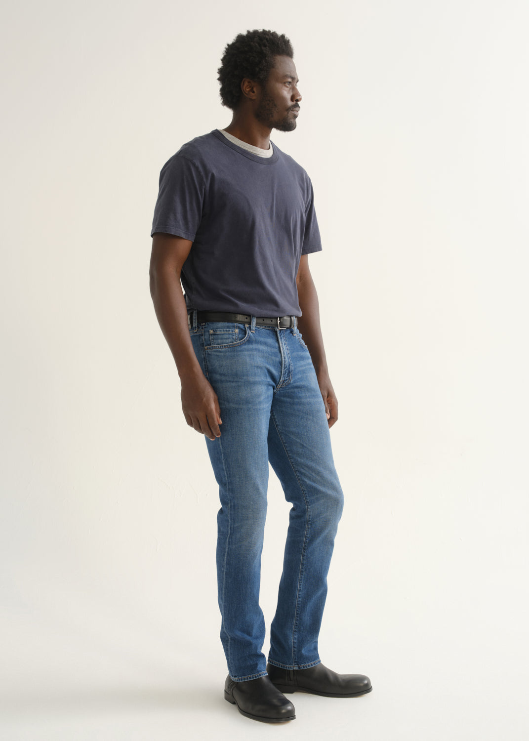 Person wearing a navy blue short sleeve crewneck t-shirt with medium blue jeans and black shoes, side full body view on a light grey background.