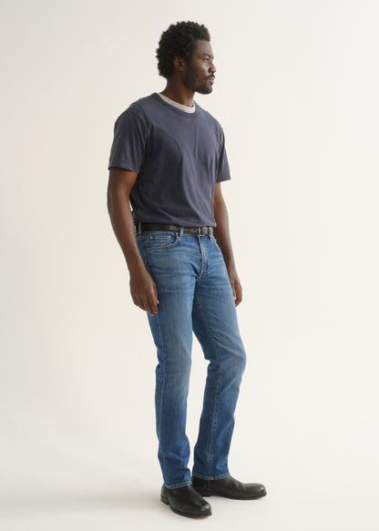 Person wearing a navy blue short sleeve crewneck t-shirt with medium blue jeans and black shoes, side full body view on a light grey background.