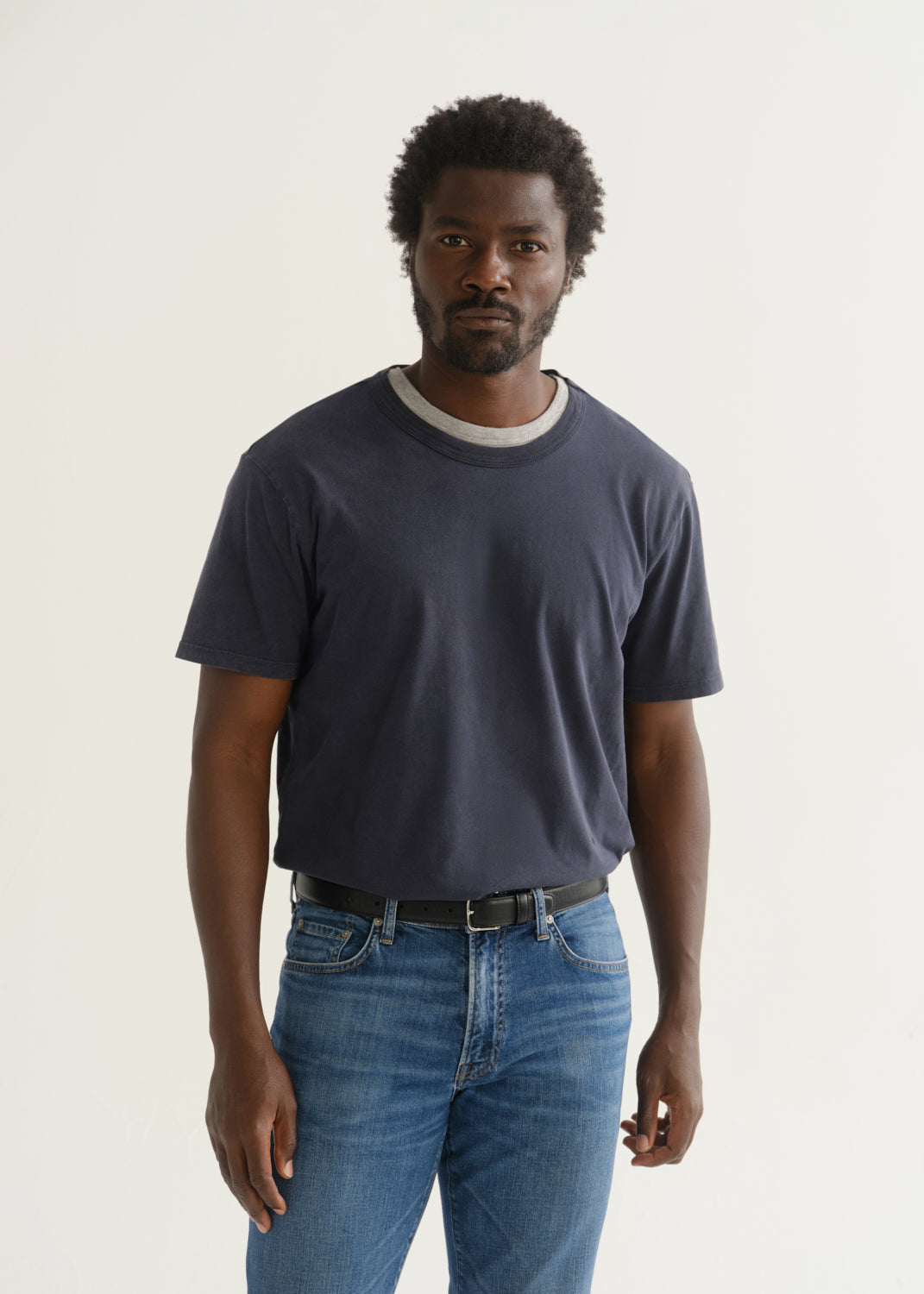 Person wearing a navy blue short sleeve crewneck t-shirt with medium blue jeans, front cropped view on a light grey background.