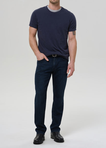 A man modeling a dark navy blue crewneck t-shirt and dark blue jeans with black leather shoes, standing against a grey background.