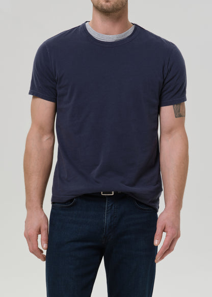 A man shown wearing a dark navy blue short-sleeve t-shirt and dark blue jeans against a grey background.