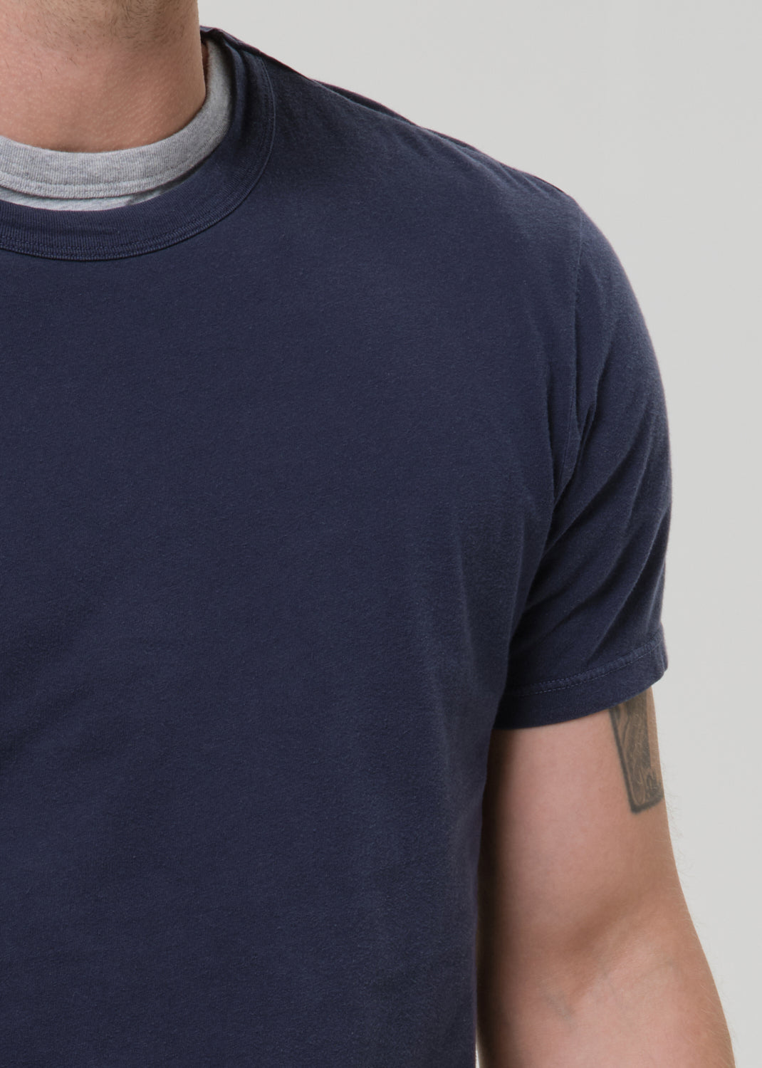 A close-up of a man wearing a dark navy blue crew-neck t-shirt over a light grey undershirt, against a grey background.