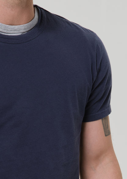 A close-up of a man wearing a dark navy blue crew-neck t-shirt over a light grey undershirt, against a grey background.