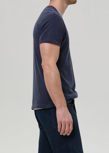 A man shown in side profile wearing a dark navy blue short-sleeve t-shirt and dark blue jeans against a grey background.