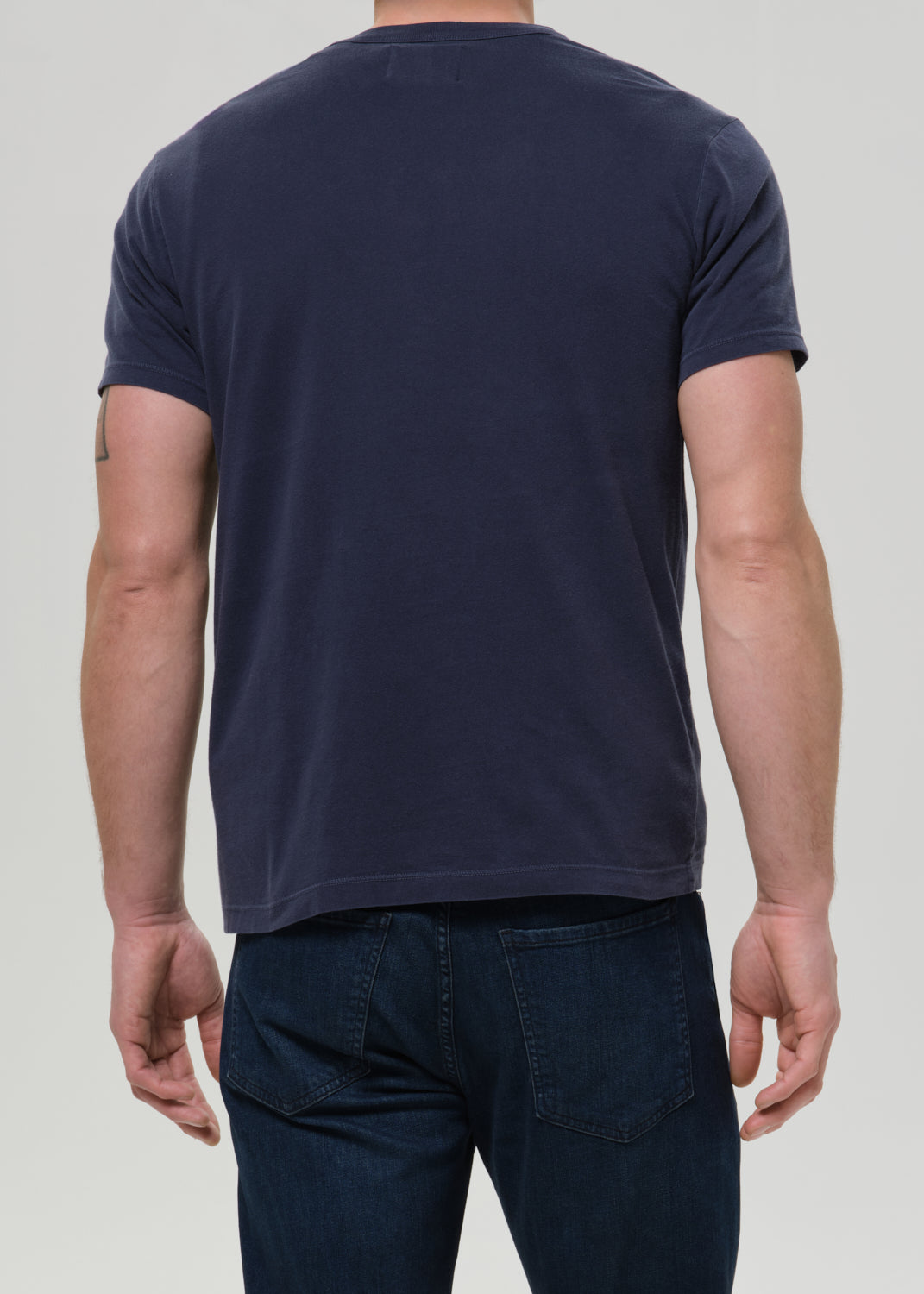 A man shown wearing a dark navy blue short-sleeve t-shirt and dark blue jeans against a grey background.